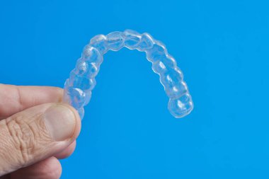 Net bir diş hizalayıcısı, şeffaf tasarımını gösteren bir el tarafından tutuluyor. Parlak mavi arkaplan hizalayıcının görünürlüğünü artırır.