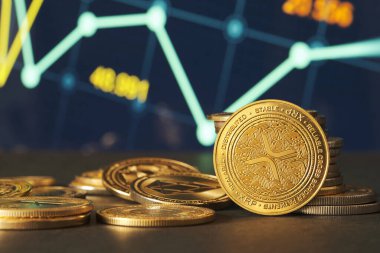 Altın XRP sikkeleri, pazar eğilimlerini ve kripto para birimlerindeki dalgalanmaları temsil eden yeşil ve mavi finansal grafikler önünde modern bir ortamda sergilenmektedir..