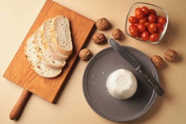 Bir kalıp peynirin yanında dilimlenmiş ekmek, taze vişneli domates ve gri tabakta bütün ceviz. Ayarlama basit bir kırsal atıştırmanın altını çiziyor..