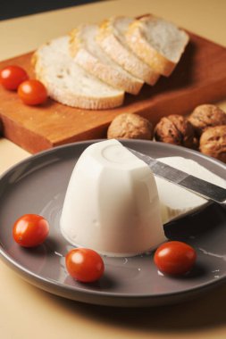 Bir kalıp peynirin yanında dilimlenmiş ekmek, taze vişneli domates ve gri tabakta bütün ceviz. Ayarlama basit bir kırsal atıştırmanın altını çiziyor..