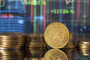 Altın XRP sikkeleri, pazar eğilimlerini ve kripto para birimlerindeki dalgalanmaları temsil eden yeşil ve mavi finansal grafikler önünde modern bir ortamda sergilenmektedir..
