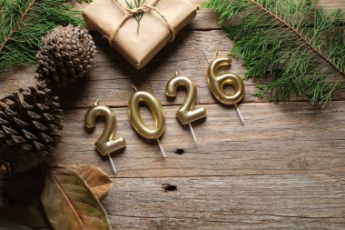 Altın mumlar 2026 'yı gösteriyor. Köhne ahşap bir yüzey üzerinde çam kozalakları ve yeşilliklerle çevrili. Yeni yıl kutlamalarının şenlikli havasını güçlendiriyor..