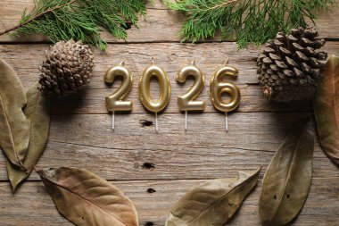 Altın mumlar 2026 'yı gösteriyor. Köhne ahşap bir yüzey üzerinde çam kozalakları ve yeşilliklerle çevrili. Yeni yıl kutlamalarının şenlikli havasını güçlendiriyor..