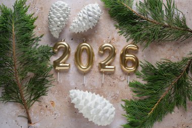 Dekoratif sergide beyaz çam kozalakları ve yeşil yapraklarla birlikte altın 2026 mum yer alarak, Yeni Yıl kutlamaları için neşeli bir ortam oluşturuldu..