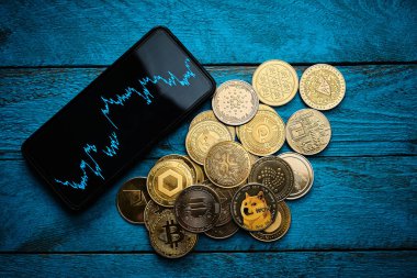 Bir akıllı telefon yükselen bir kripto para grafiğini gösterirken çeşitli paralar mavi ahşap bir masaya saçılmıştır. Sahne dijital finans eğilimlerini vurguluyor.