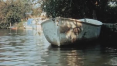 A serene view from a sailboat as it glides down a tranquil river surrounded by lush greenery. Rustic boats docked alongside the river bank. Longridge Activity Centre, River Thames. Historical footage