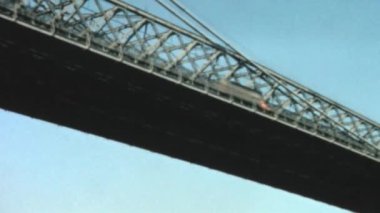 Low-angle view of the Brooklyn Bridge in New York City. Cars and other vehicles drive along it. The bridges intricate structure is fully displayed, with its suspension cables. Archival footage of