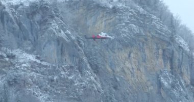 Helicopter ascends vertically with a load of logs hanging from a rope. Gray, cloudy sky on a winter morning with bluish light. Snowy mountain as background. Kaman K-MAX