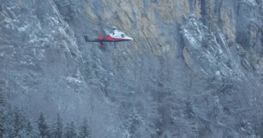 Helicopter ascends vertically near the slope of a snow covered mountain with a hanging rope on a grey, cloudy winter morning. Kaman K-MAX. Sense of excitement and admiration for the skill and