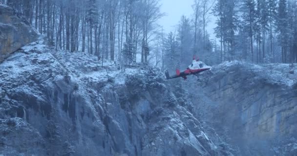 Вертолет Медленно Спускается Вертикально Скалистого Склона Снежной Горы Сосновым Лесом — стоковое видео