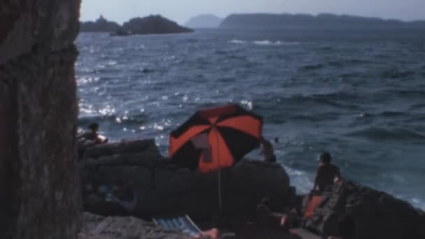 この1970年のクリップは ドゥブロヴニクのビーチがどれだけ活気があったかを示しています 美しい景色は明るい日差し 穏やかな波 そしてビーチにいる間に日焼けしたカップルによって設定されています パラソルの移動 — ストック動画