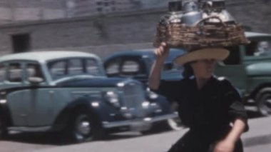 Kafasında sepet ve metal kaplarla yürüyen beyaz bir kadın var. 1950 'lerin arabaları arka planda park edilmiş. Santa Cruz de Tenerife Arşivi 'ndeki Nuestra Senora de Africa Pazarı.