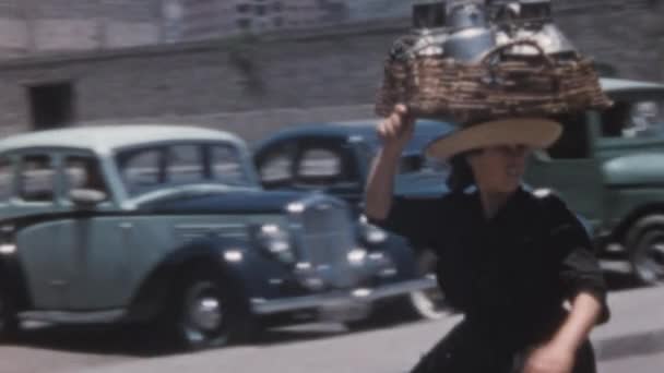若い白人女性は彼女の頭の上にバスケットと金属容器で歩く 1950年代の車両が背景に停まっていた サンタ クルス テネリフェのヌエストラ セノーラ アフリカ市場アーカイブビデオ — ストック動画