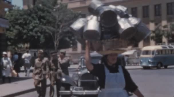 女人头上挂着很多金属容器走路 20世纪50年代的汽车停放在背景中 Santa Cruz Tenerife Nuestra Senora Africa Market回顾桑尼日的怀旧镜头 — 图库视频影像