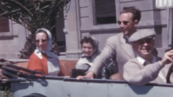 1950年代 在西班牙大加那利亚 坐在老式灰色轿车上的妇女和男子 女人头上戴着围巾 戴着太阳镜 在一个阳光灿烂 没有汽车的炎热夏天 男人们都戴着一顶帽子和一套衣服 — 图库视频影像