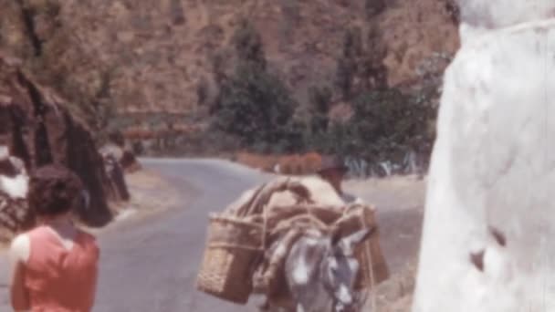 帽子を被った老人が暑い夏の日に道路を歩いていて 1950年代にグラン カナリア スペインで大きな灰色の岩を通過しています 男性は暗いカバーとログスリーブを着ています暖かい晴れた日に — ストック動画