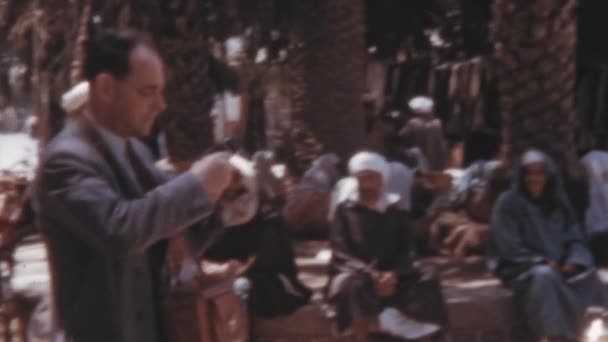 地元の人々が衣類や台所用品を販売している小さな地域で道端の市場の写真を撮る男性観光客 1950年のモロッコ ジャディダの歴史的映像 — ストック動画