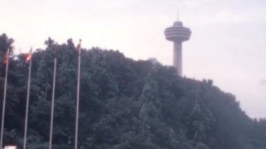 1970 'lerin arşiv görüntülerinde Niagara Şelalesi Ontario Kanada' sında bayrak direğinin tepesinde Amerikan ve Kanada bayrakları dalgalanıyor. Skylon Kulesi arka planda, bir tepenin üstünde yükseliyor. Proje için mükemmel