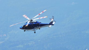 Sloven polisinin mavi Leonardo AW169 helikopteri güneşli bir günde yemyeşil tepelerin üzerinde uçuyor. Boşluğu kopyala.