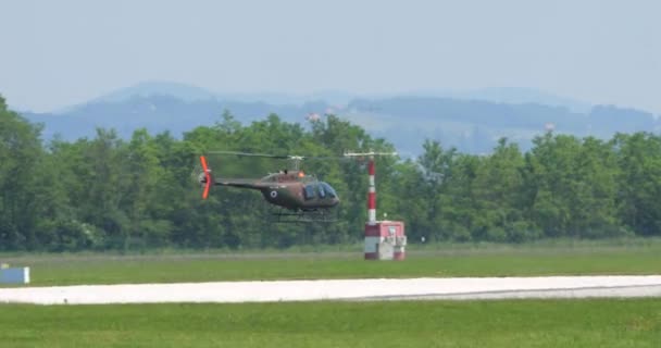 2023年6月4日 一架绿色伪装的贝尔206型军用直升机在机场跑道上空低空飞行 另一架直升机停放 — 图库视频影像