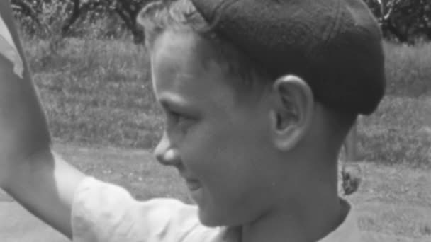 少年は木と草のあるオープンエリアで夏の太陽を楽しんでいます 友達が遠くで遊んでいるのを見ながら笑顔 クラシックキャップを着用 彼の目には無実だった 1930年代 ニューヨーク ヴィンテージ フッテージ — ストック動画