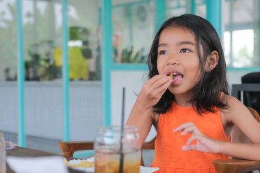 Asyalı anaokulu kızı tek başına oturuyor ve mutlu bir şekilde patates kızartması yiyor.