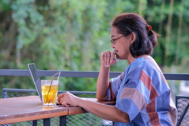 Asyalı bir kadın evinin balkonunda oturmuş, dizüstü bilgisayarıyla masada bir bardak limon çayıyla çalışıyor.