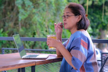 Ev terasında dizüstü bilgisayarı üzerinde çalışırken buzlu limon çayı içen Asyalı kadın.