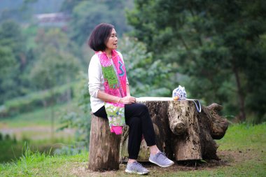 Asyalı orta yaşlı kadın bir kütüğün üzerinde tek başına oturuyor, kırsalda güneşleniyor.