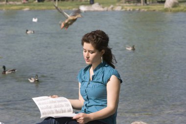 Kız sessizce gölün kenarında bir şey okuyor. Dalgalı saçlı, mavi bluzlu, su kenarında kitap okuyan bir kız..