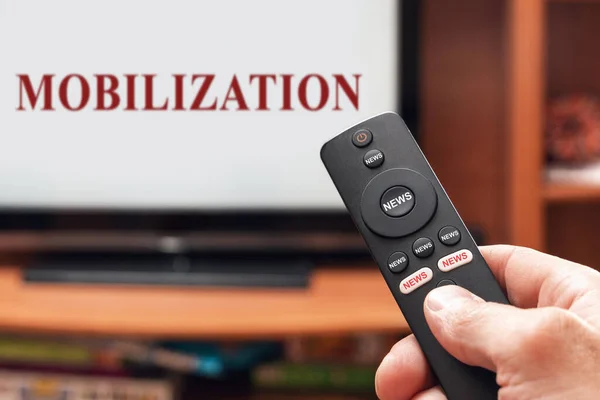 A man\'s hand holds a TV remote control, all buttons on the remote control are signed news, on the TV there is an inscription mobilization.