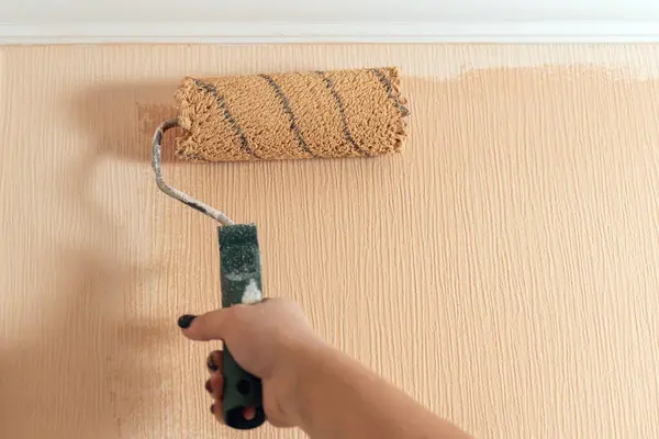 A woman\'s hand paints a wall with a paint roller in a beige color.