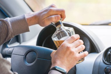 Şoför direksiyonda oturuyor ve elinde bir şişe alkollü içecek tutuyor. Kanunları çiğnemek ve araba kullanırken alkol içmek..