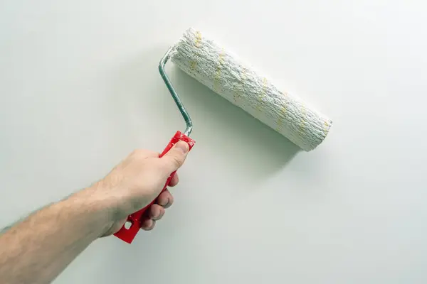 A man\'s hand paints a wall white with a paint roller.