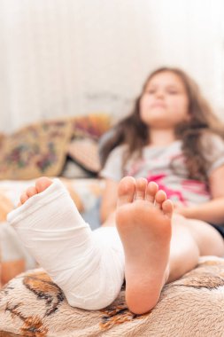 Kırık bacaklı, alçılı küçük kız yatakta oturuyor..