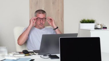 Yorgun görünüşlü bir adam dizüstü bilgisayarla çalıştıktan sonra gözlüğünü çıkarır.