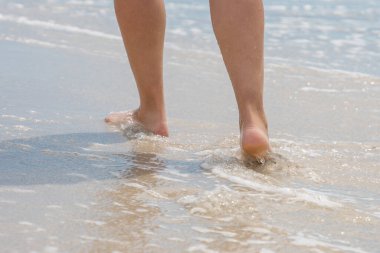 Güneşli bir yaz gününde kumlu bir sahil boyunca yürüyen bir kız, deniz suyu ayaklarını yıkar..