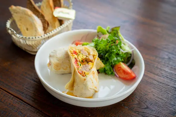 Tabakta yeşil yapraklar, domates dilimleri ve sebzeler içeren çeşitli malzemelerle dolu rulo tortilla.