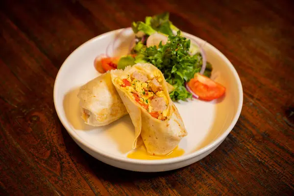 Tabakta yeşil yapraklar, domates dilimleri ve sebzeler içeren çeşitli malzemelerle dolu rulo tortilla.
