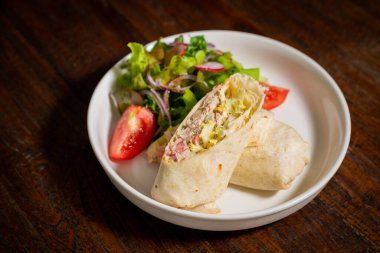 Tabakta yeşil yapraklar, domates dilimleri ve sebzeler içeren çeşitli malzemelerle dolu rulo tortilla.