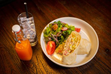 Kahvaltı tabakta, yanında tortilla ve salata ile. Burrito kısmen açık et ve sebzeler dahil, arka plan ise ahşap bir masa.