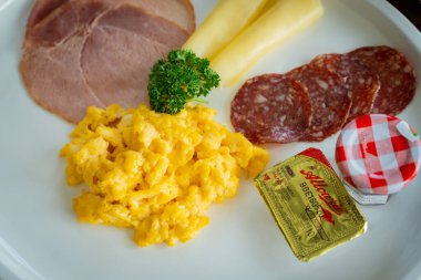 Bir tabağa yayılmış kahvaltıyı kapat, çırpılmış yumurta, jambon, salam, peynir çubuğu ve küçük bir kavanoz reçel dahil.