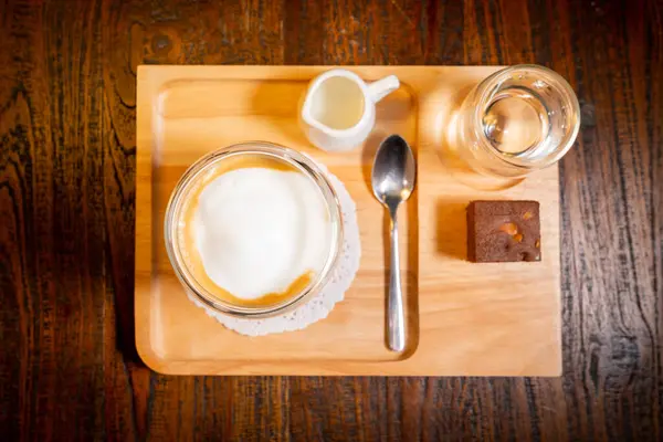 Bir bardak mocha kahvenin üst görüntüsü, küçük bir şurup sürahisi, bir bardak su, kaşık ve ahşap tepside bir brownie.