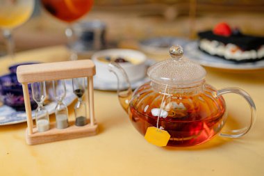 Çay dolu cam çaydanlık ve yanında asılı sarı etiket, ahşap kum saati.