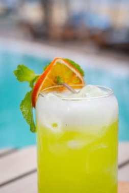 Close up view, Midori Sour cocktail with slice dried lemon and mint leaves on wooden planks in front of a pool clipart