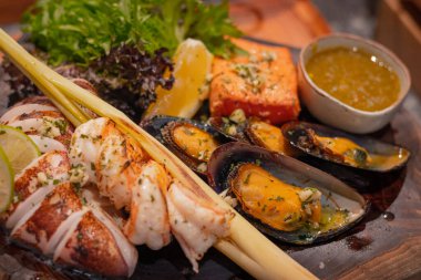 Yakın mesafede, ızgara deniz ürünleri kalamar, somon, midye, Tayland bitkisi ve küçük bir kase ahşap tabakta deniz ürünleri sosu bulunur.