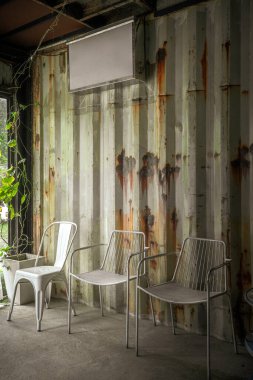 İçinde üç beyaz metal sandalye bulunan metal bir oda yıpranmış, paslı bir metal duvarın önünde.