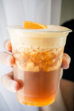 Yakın plan çekimde, şeffaf bir fincan dolusu köpüklü kremalı ve turuncu dilimli kahve bardağını kenarda tutarken.