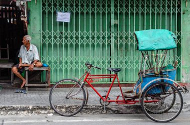 Chon Buri, Tayland - 8 Aralık 2018: Yeşil bir kapının yanındaki bankta oturan yaşlı bir adam, metal ızgaralı, mavi kenarlı kırmızı bir bisiklet, yeşil bir branda ile kaplanmış.