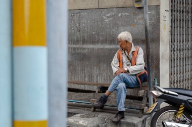 Chon Buri, Tayland - 9 Aralık 2018: Yaşlı bir adam tahta bir bankta beton bir duvara karşı oturur, beyaz gömlek, turuncu yelek ve mavi kot pantolon giyer.